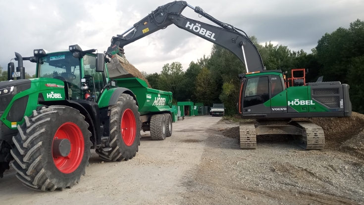 Fendt Vario mit Muldenkipper beim Materialtransport auf einer Baustelle der Ernst Höbel GmbH. Ein Kettenbagger belädt den Anhänger mit Schüttgut für den weiteren Transport.