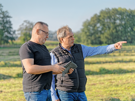 Andreas Osters and Christof Voß are standing on a meadow. The one on the left is holding a tablet and looking at the screen. The one on the right is pointing straight ahead with his right hand.