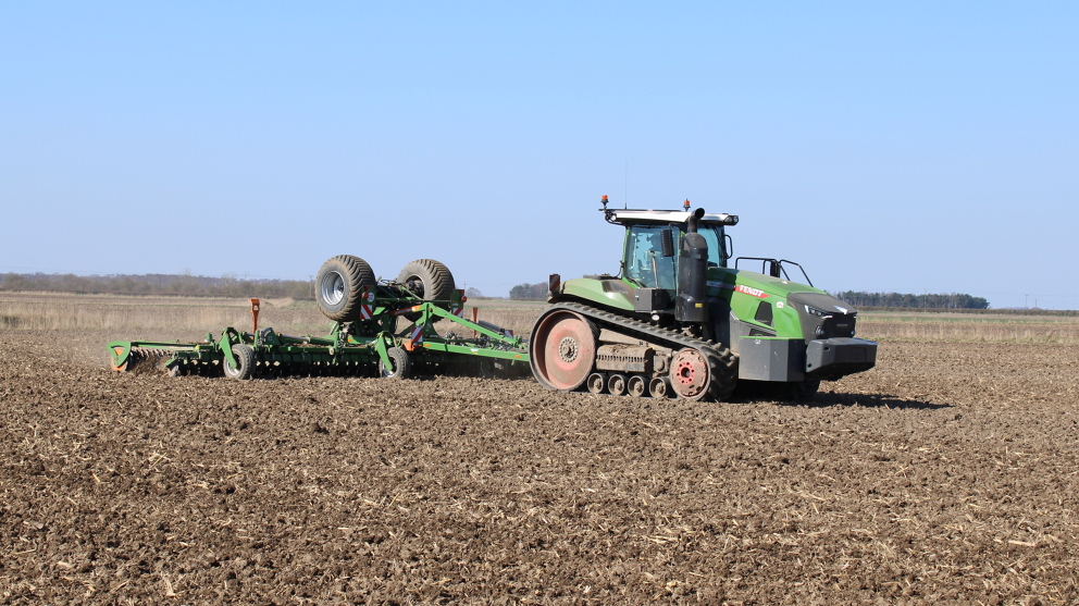 A picture from the front side of the Fendt 1167 Vario MT pulling a green drill across a field with a clear blue sky in the background