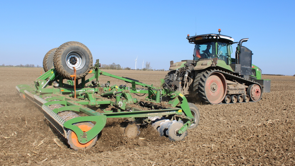 A picture from the backhand side of the Fendt 1167 Vario MT pulling a drill across a field with some wind turbines in the background and a clear blue sky