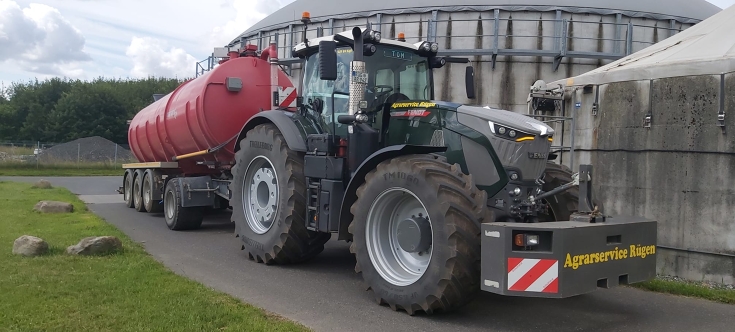 Pine green Fendt 900 Vario parked with a red slurry tanker in front of a large slurry storage tank. The tractor is used by Agrarservice Rügen for efficient slurry application and nutrient management.