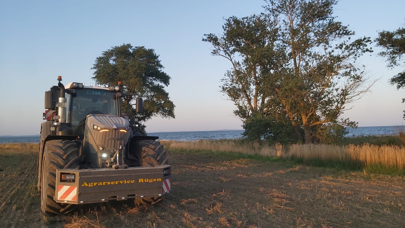 Pine green Fendt 900 Vario standing on a harvested stubble field on the island of Rügen, with the Baltic Sea visible in the background. The tractor features Agrarservice Rügen’s distinctive dark green colour scheme.