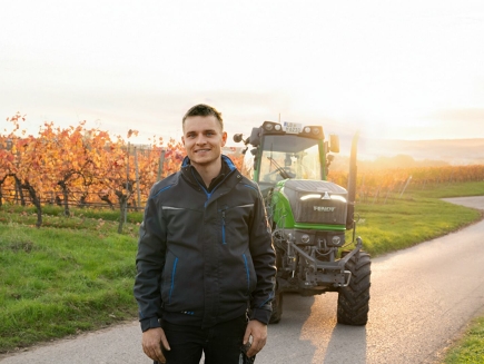 Tobias Händel se tient devant son tracteur spécialisé Fendt dans les vignes. En arrière-plan, on peut voir les vignes.